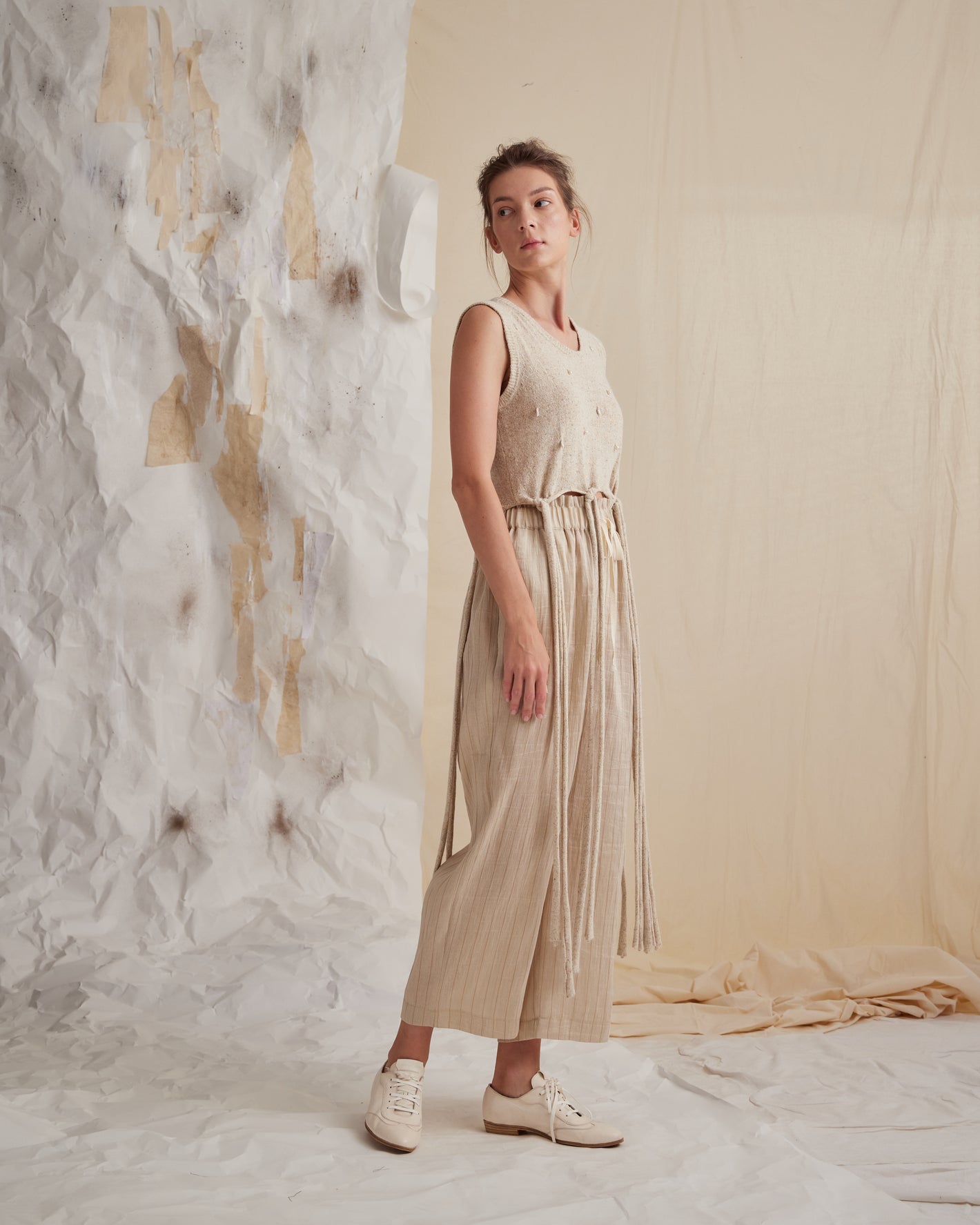 A Tentative Atelier SS23 Lookbook Womens Beige subtly striped trousers and beige knitted sleeveless top with long tied hem and pearl pendant detailing