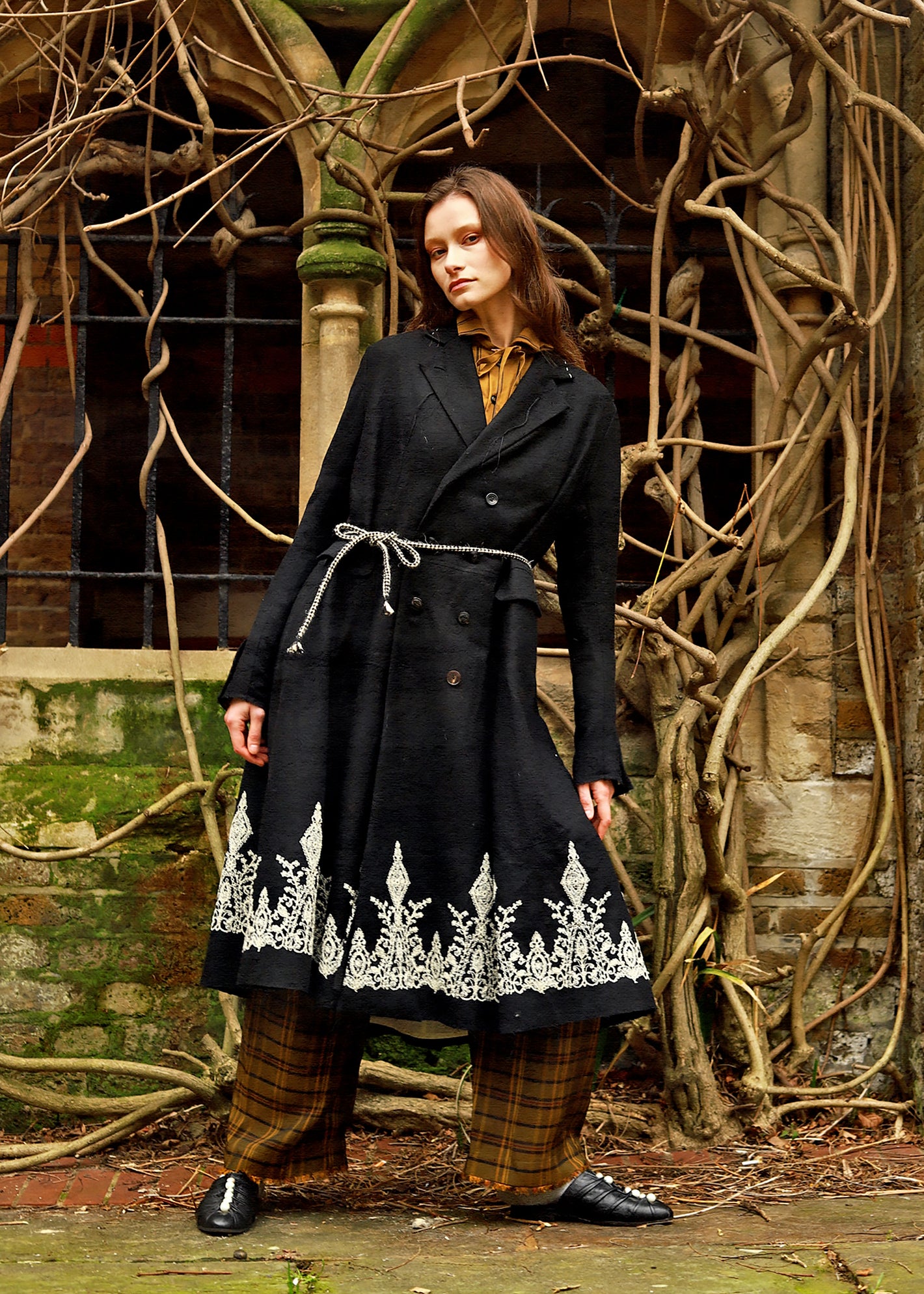 A Tentative Atelier AW23 Lookbook Womens black double breasted coat with a tie waist and embroidered hem, with brown checked trousers ensemble