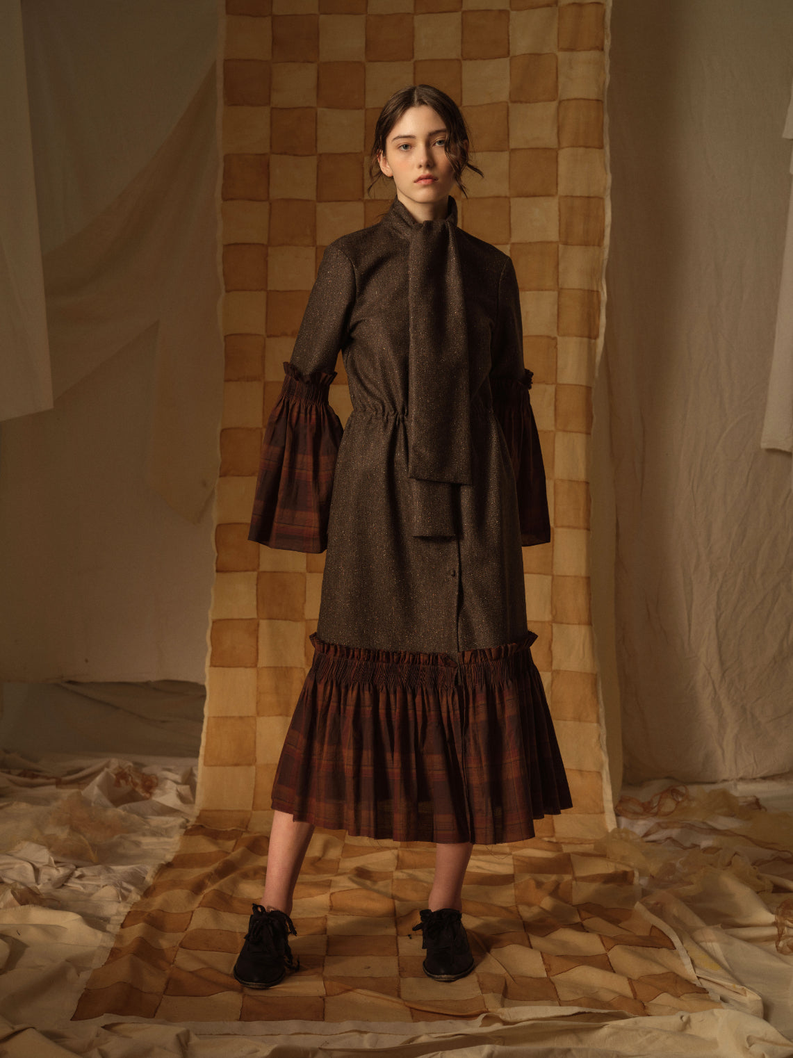 A Tentative Atelier AW21 Lookbook Womens brown wool coat with a gathered waist, scarf neck tie, and contrasting brown check ruffle hem and sleeves