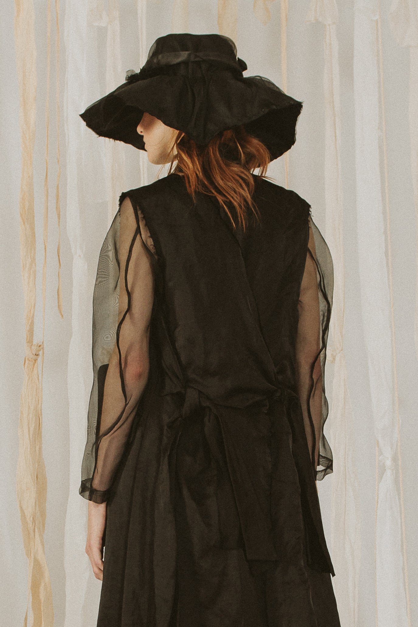 A Tentative Atelier SS18 Lookbook Womens crop of the back of black silk coat with sheer sleeves, styled with a broad rim black hat