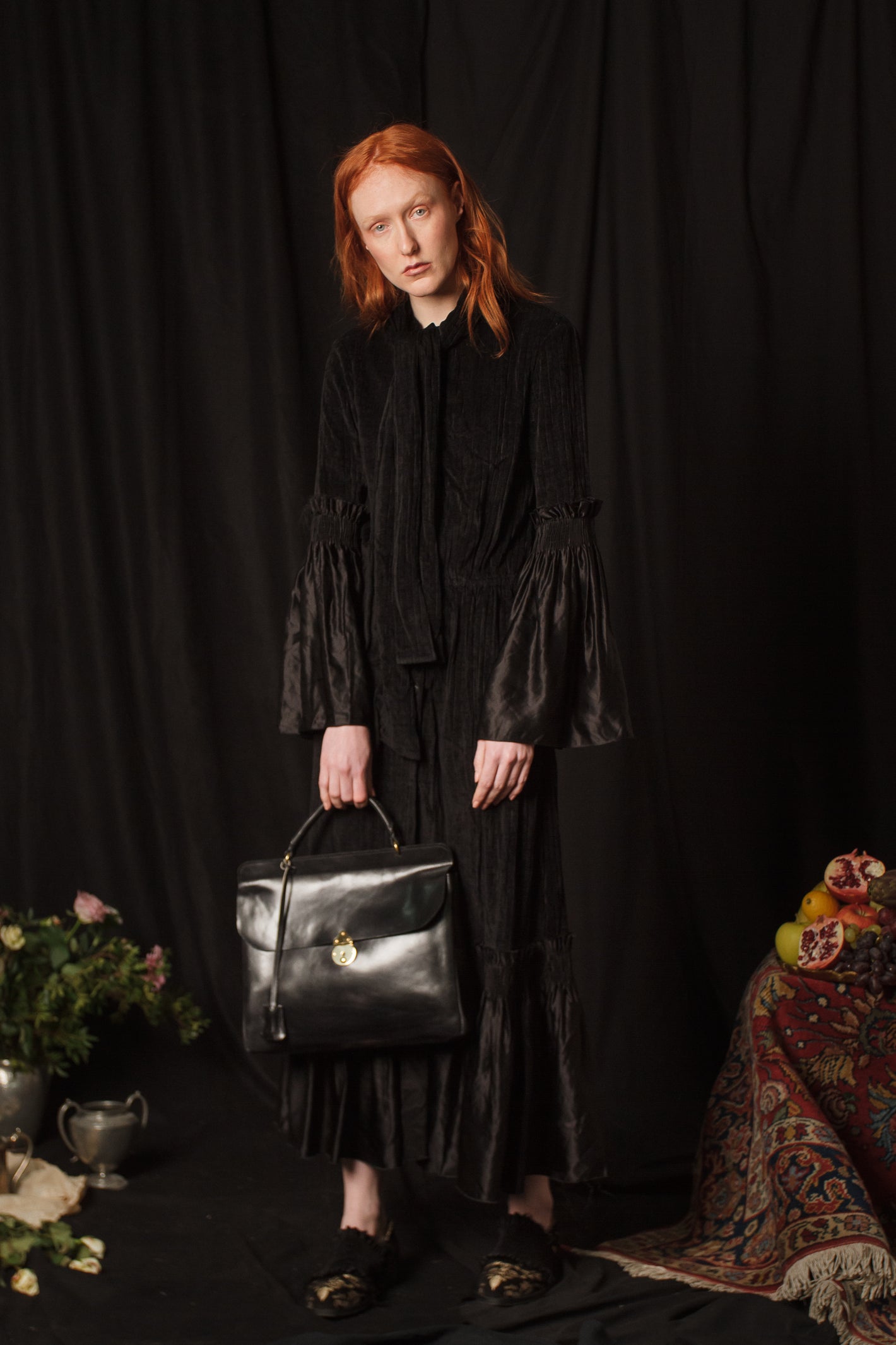 A Tentative Atelier AW18 Lookbook Womens long black dress with neck tie, contrasting black silk bell sleeves and black silk hem, styled with a lack leather top handle bag